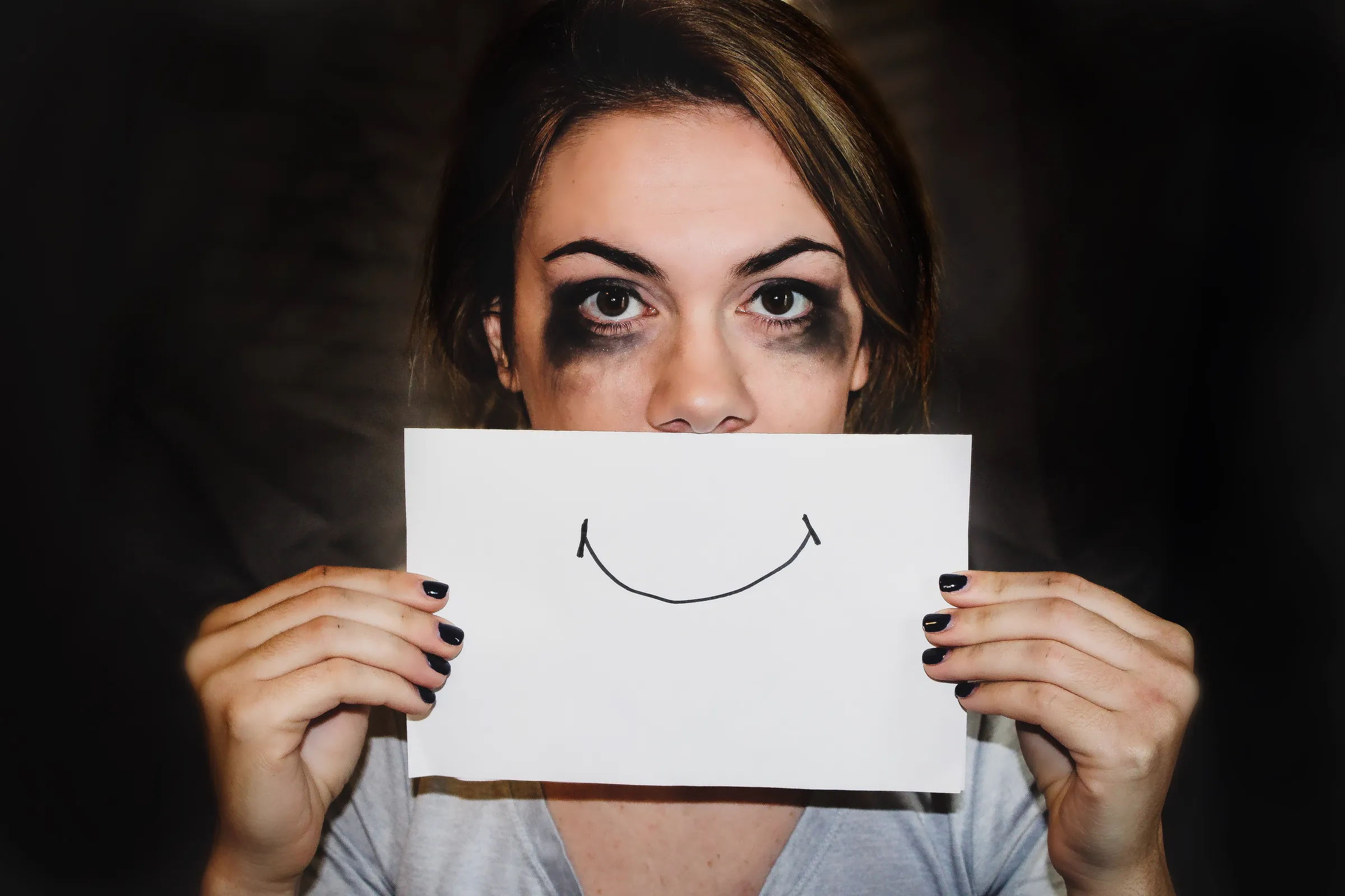 A woman with smudged mascara holding a drawn smile in front of her mouth